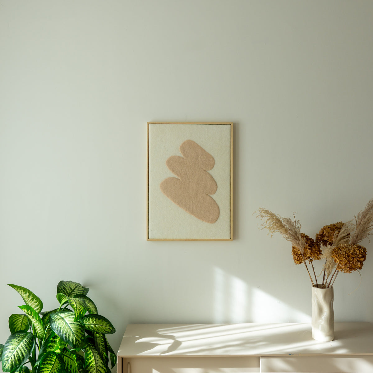 Vilten Stone wandkleed aan de muur boven een licht dressoir met plant en droogbloemen.